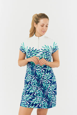 The Pure Golf close up model shot of the Carly Cap Sleeve dress showcases an aquatic design in shades of blue and ocean hues, featuring a print of floating palms from the chest down. This longer-length dress is crafted from 4-way stretch fabric for enhanced flexibility, with detachable shorts for added coverage. Its flattering fit suits all body types, while the mock neck collar delivers a sleek style. Designed to resist creasing, static, and moisture, the dress also offers UPF 50+ protection.